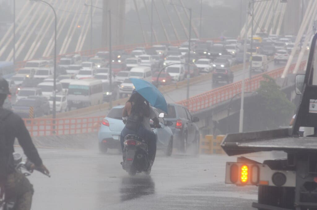 Una mujer, en la parte trasera de una motocicleta, se protege con una sombrilla esta mañana de las lluvias que caían sobre gran parte del Gran Santo Domingo, mientras al fondo se puede ver a hilera de vehículos y la neblina sobre el puente Juan Bosch. Según Meteorología, las lluvias son provocadas por una vaguada e incidirán sobre el territorio nacional durante las próximas 48 horas./Foto Jorge González